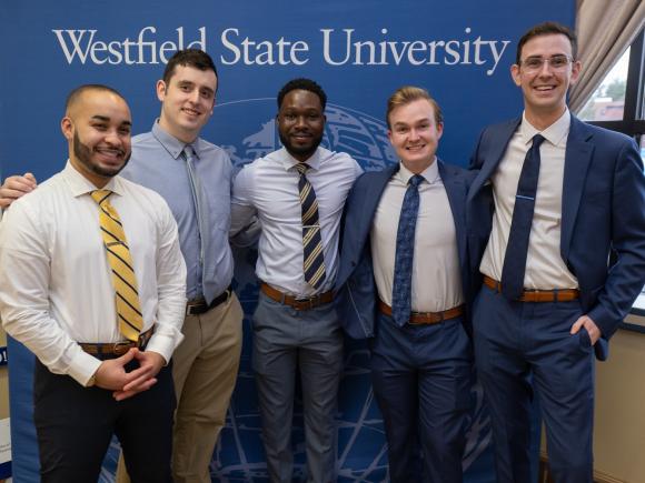 Graduate students in professional attire smiling in front of a university banner.