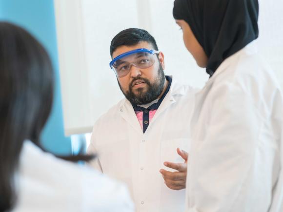 Roderico Acevedo teaches in a lab coat and goggles while two students also in lab coats look on